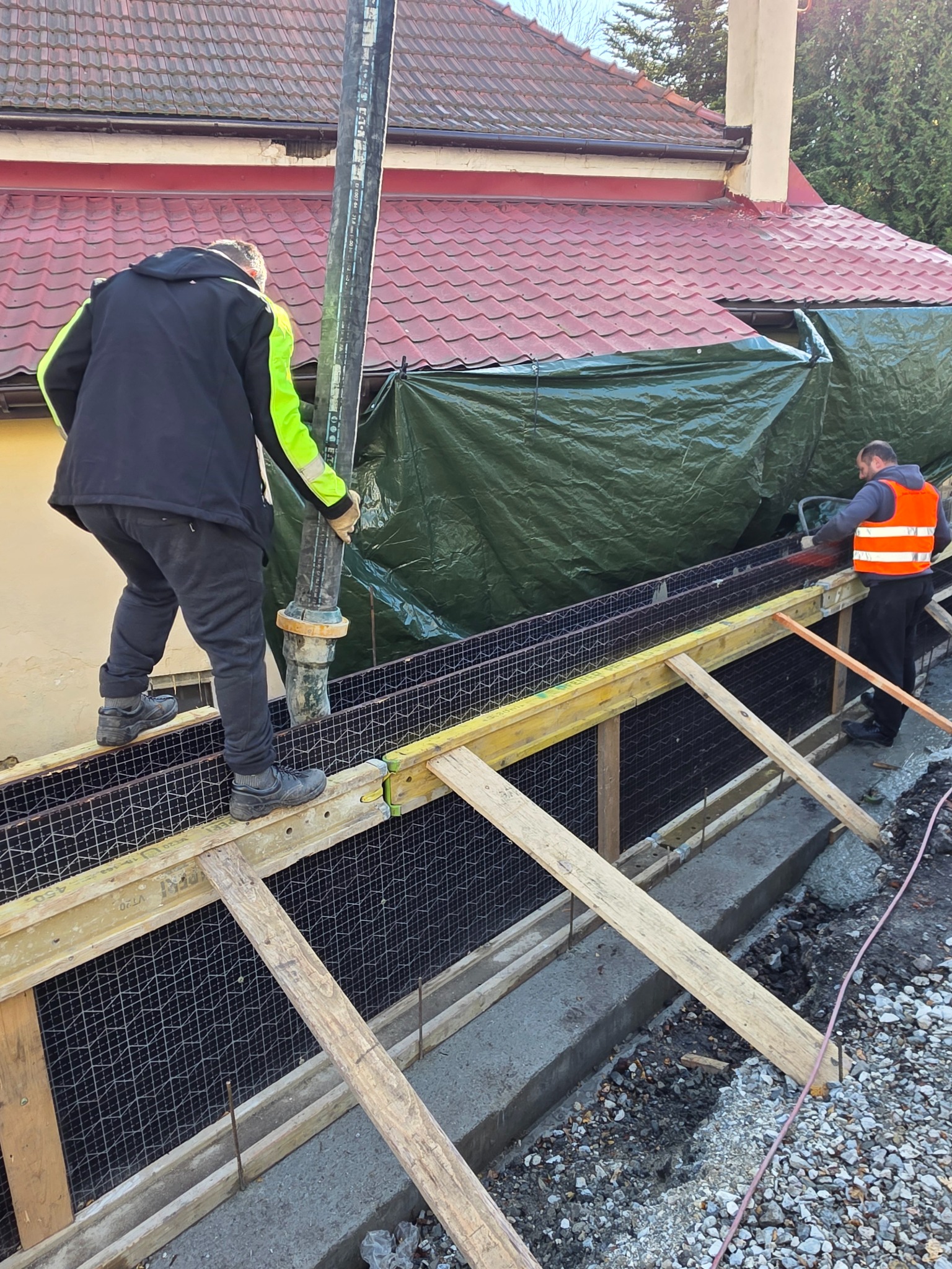 Wylewka betonu do szalunku zbrojonego siatką, widoczna pompa do betonu i pracownicy w odzieży roboczej. W tle budynek z czerwonym dachem i zielona plandeka.