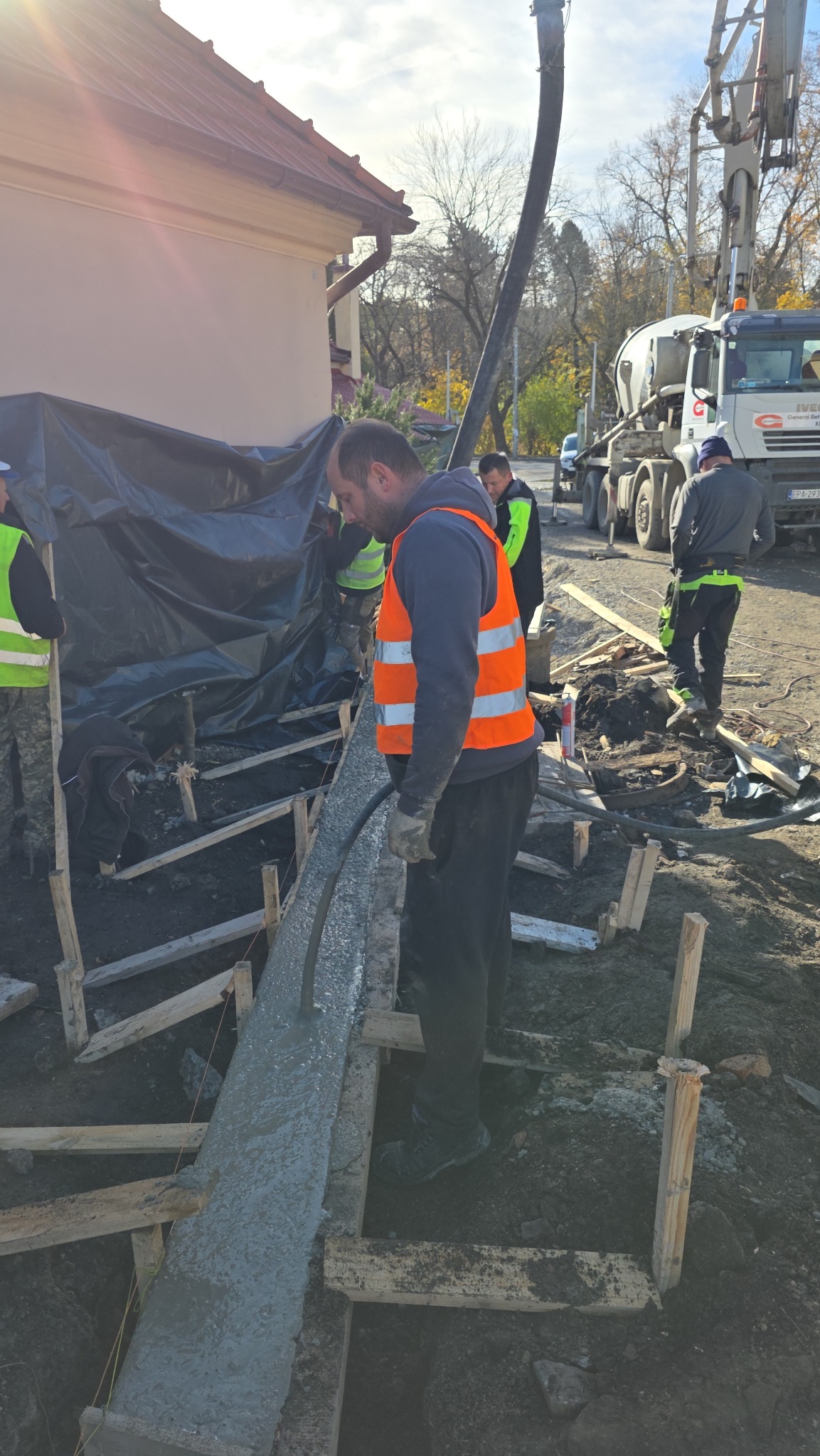 Wylewanie betonu do szalunku pod fundament. Pracownicy w kamizelkach odblaskowych, pompa do betonu w tle. Etap budowy domu jednorodzinnego.