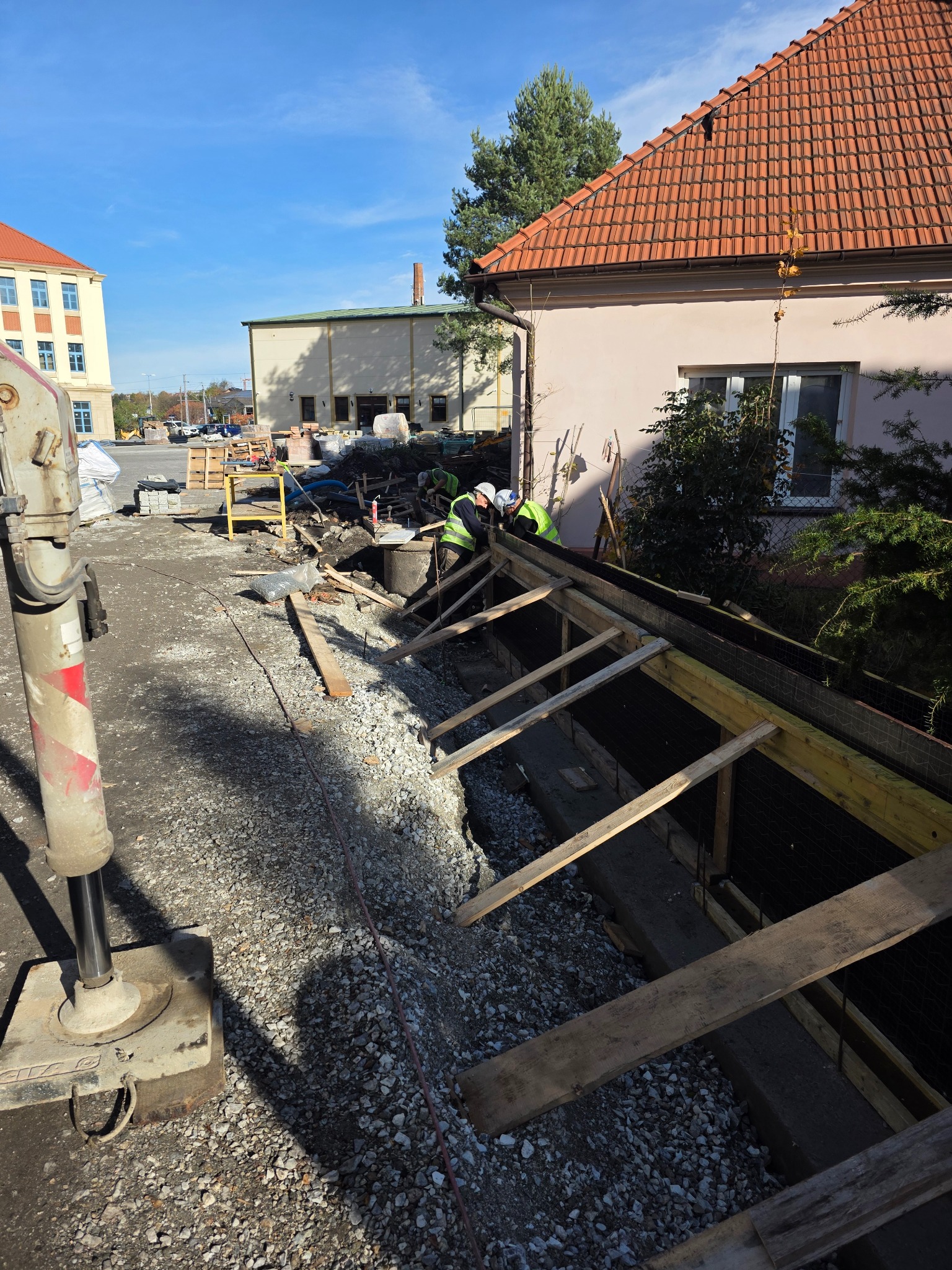 Budowa fundamentów: szalunek z drewnianych belek, widoczni pracownicy w kaskach i kamizelkach odblaskowych, gruz i maszyna budowlana w tle, słoneczny dzień.