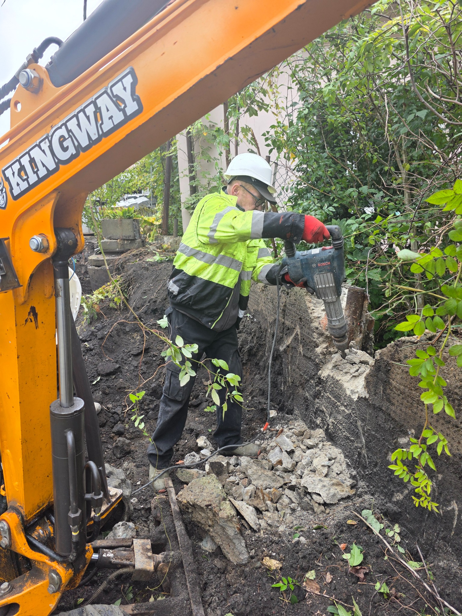 Pracownik w kasku i kamizelce używa młota pneumatycznego do rozbiórki fundamentów, obok widoczny fragment pomarańczowej koparki Kingway.