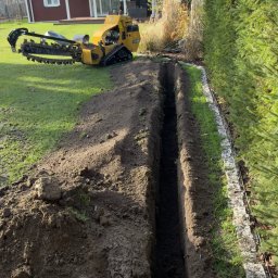 PRACE W ZIELENI SPÓŁKA Z OGRANICZONĄ ODPOWIEDZIALNOŚCIĄ - Wąski wykop w trawniku, wykonany specjalistyczną maszyną. W tle dom z czerwonej cegły i zielony żywopłot. Wykop przygotowany pod instalację.