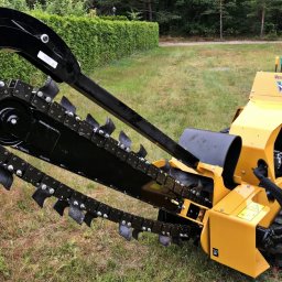 PRACE W ZIELENI SPÓŁKA Z OGRANICZONĄ ODPOWIEDZIALNOŚCIĄ - Żółta frezarka łańcuchowa Vermeer RT250 na gąsienicach, z czarnym ramieniem i łańcuchem tnącym, na trawiastym terenie z żywopłotem w tle.
