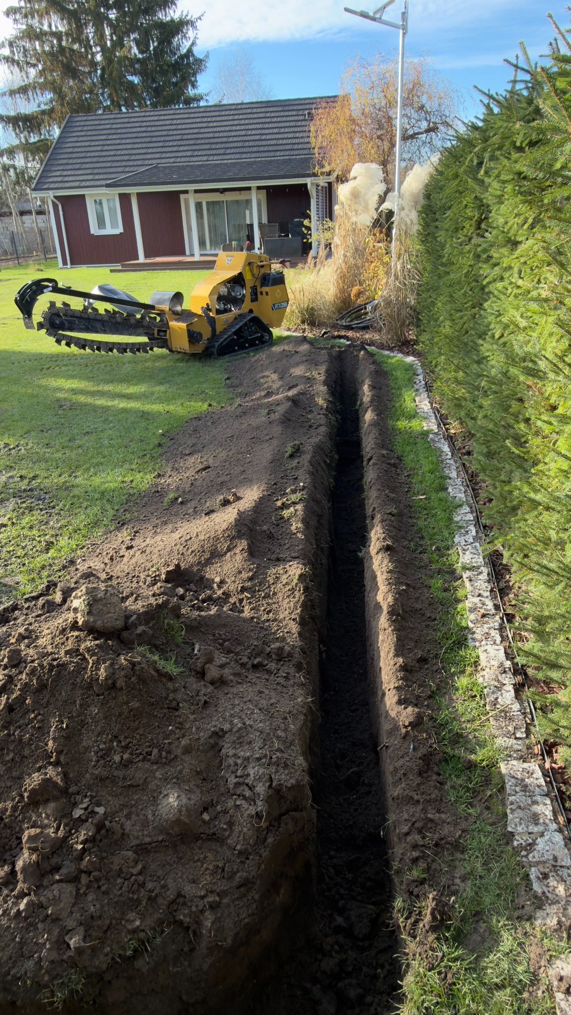 Wąski wykop w trawniku, wykonany specjalistyczną maszyną. W tle dom z czerwonej cegły i zielony żywopłot. Wykop przygotowany pod instalację.