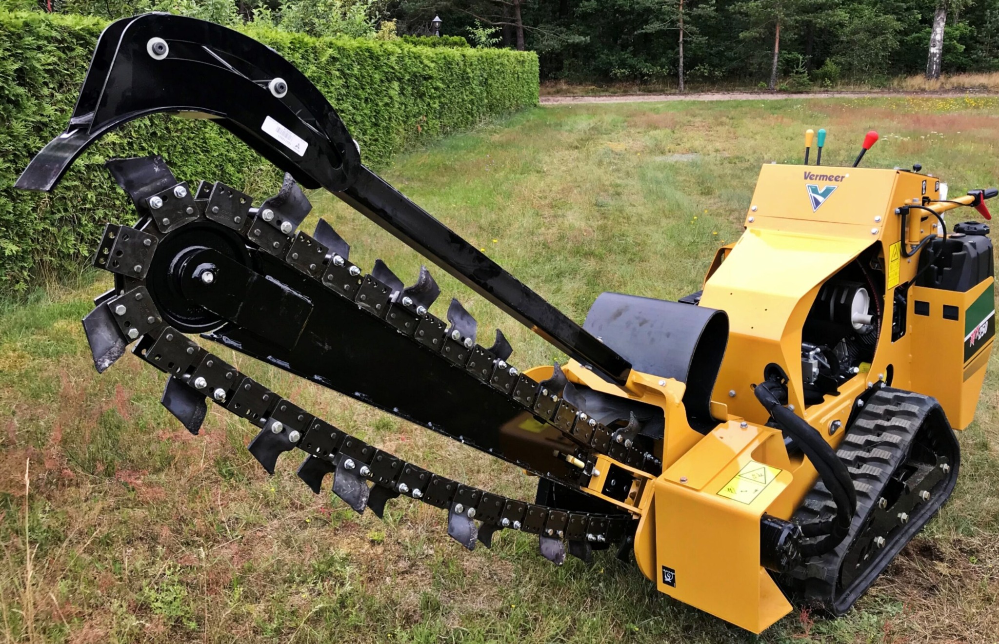 Żółta frezarka łańcuchowa Vermeer RT250 na gąsienicach, z czarnym ramieniem i łańcuchem tnącym, na trawiastym terenie z żywopłotem w tle.