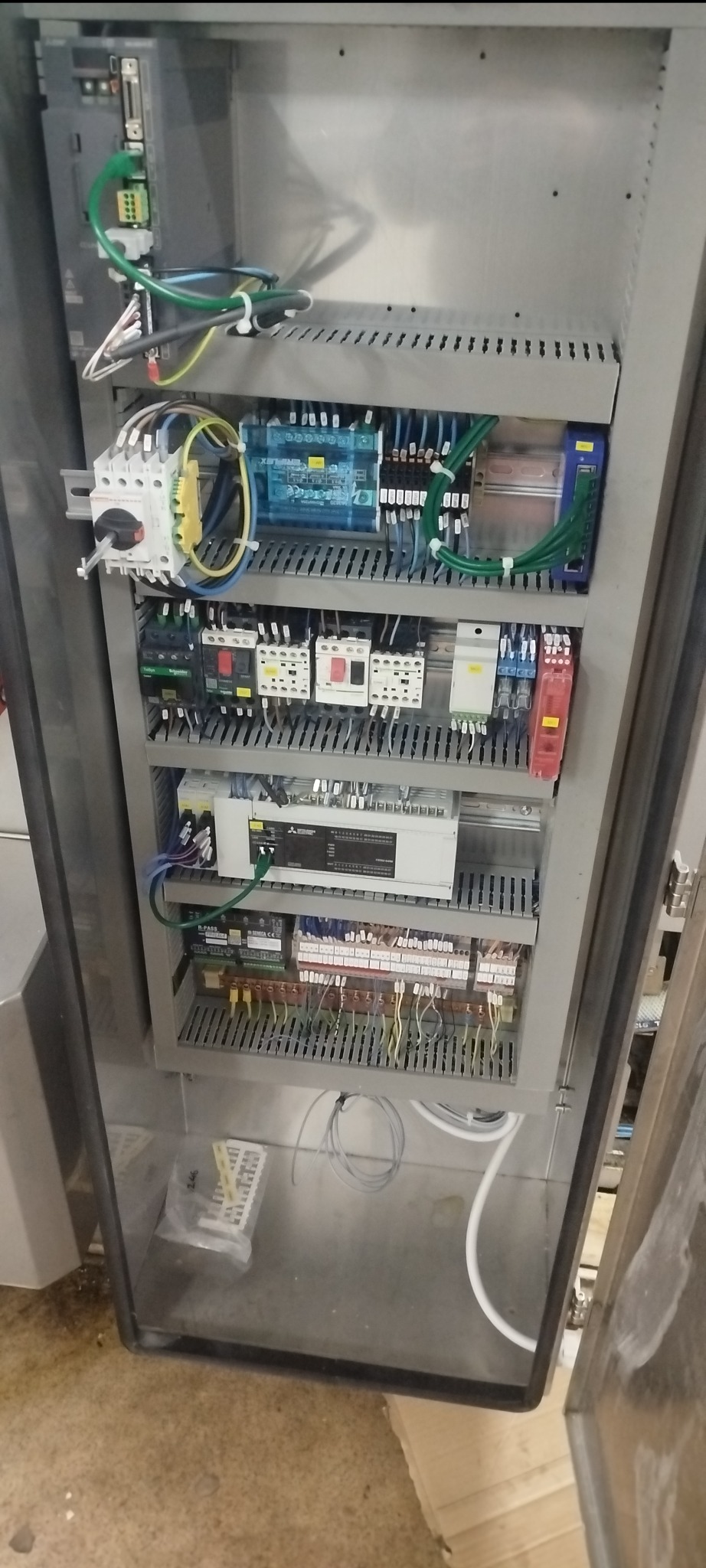 Rozdzielnica elektryczna w metalowej obudowie z widocznymi elementami sterującymi, przewodami i zabezpieczeniami. Widoczne komponenty Mitsubishi.