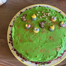 Feel A Verve Sp.z o.o. - Okrągły tort z zielonym kremem, dekorowany czekoladowymi zajączkami, kurczaczkami, jajkami i złotymi kuleczkami. Brzeg tortu obsypany płatkami róż. Całość na białym talerzu z żółtym obramowaniem.