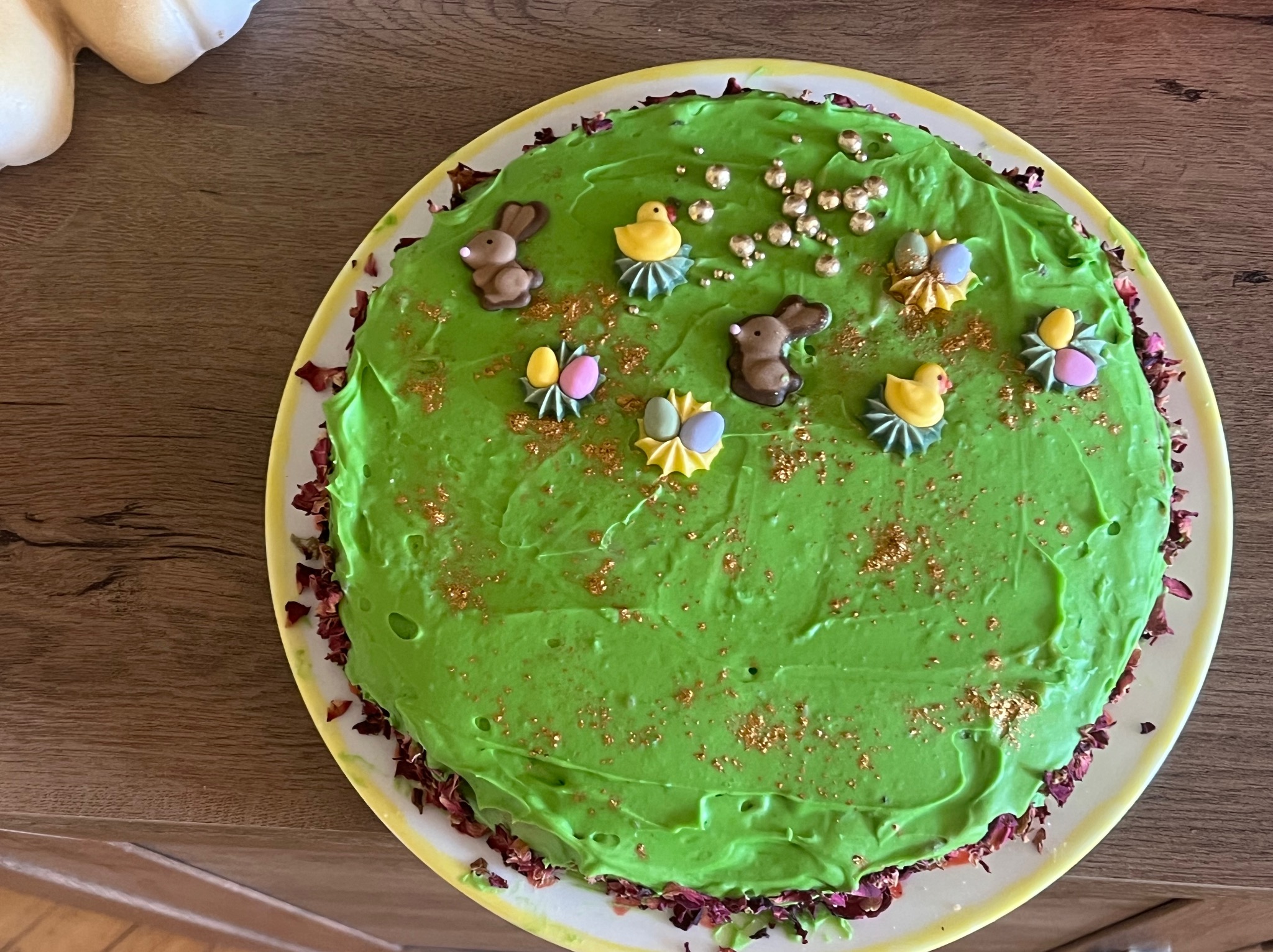Okrągły tort z zielonym kremem, dekorowany czekoladowymi zajączkami, kurczaczkami, jajkami i złotymi kuleczkami. Brzeg tortu obsypany płatkami róż. Całość na białym talerzu z żółtym obramowaniem.
