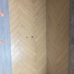 Marcin - Ściana wyłożona jasnymi płytkami w jodełkę, z widocznymi otworami na gniazdka i fragmentem szarej płytki po lewej stronie. Surowy sufit i podłoga w tle.