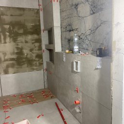 Marcin - Wnętrze łazienki w trakcie prac. Częściowo ułożone szare płytki imitujące marmur na ścianach, poziomica, wiadro z akcesoriami do układania płytek, fragment podłogi z płytkami.