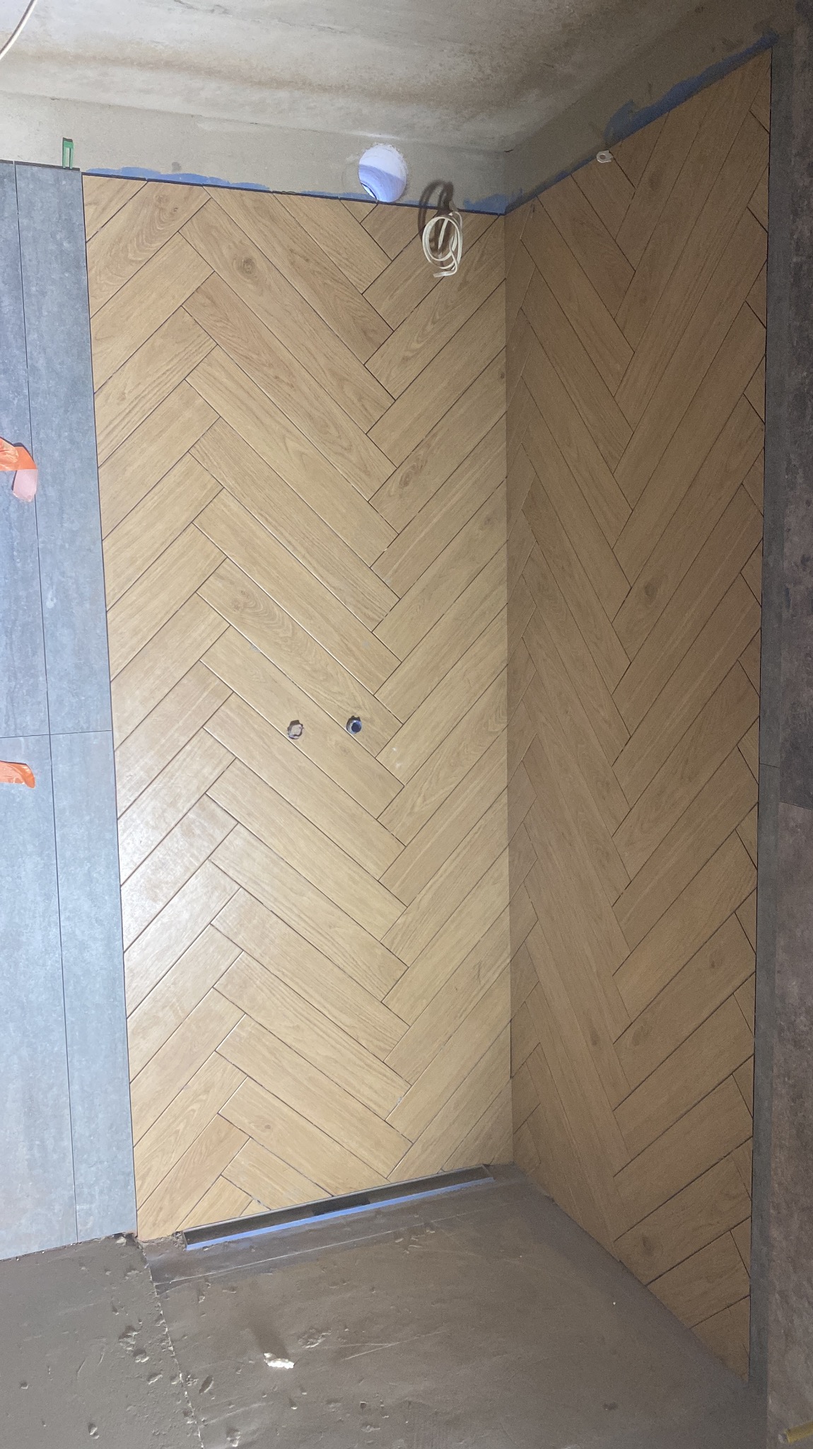 Ściana wyłożona jasnymi płytkami w jodełkę, z widocznymi otworami na gniazdka i fragmentem szarej płytki po lewej stronie. Surowy sufit i podłoga w tle.