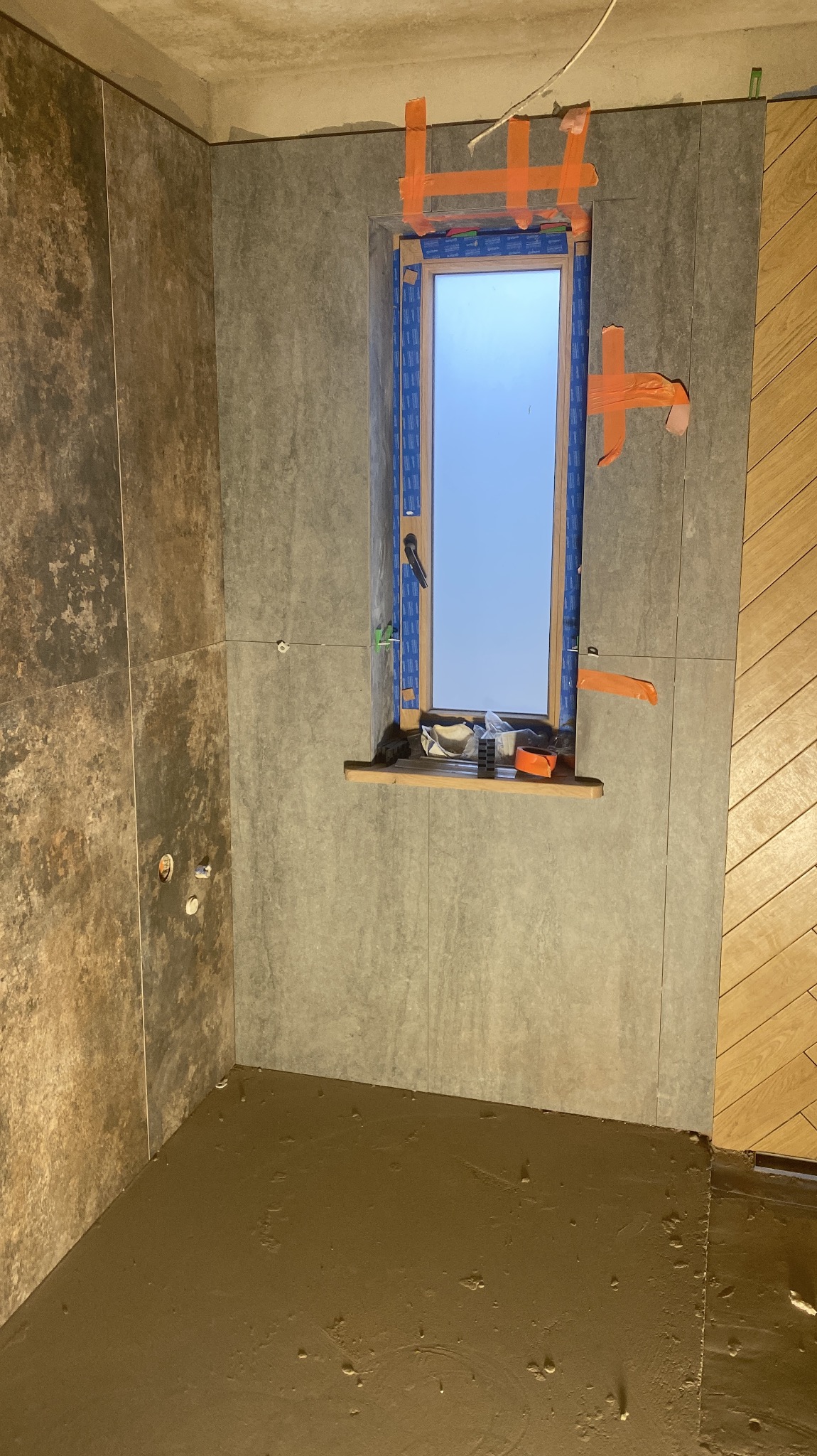 Ściany w łazience wyłożone szarymi płytkami imitującymi kamień, okno zabezpieczone taśmą, podłoga z wylewką. Widoczne narzędzia glazurnicze.