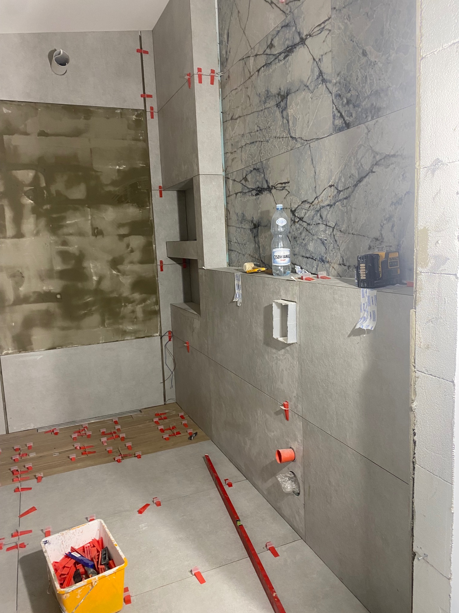 Wnętrze łazienki w trakcie prac. Częściowo ułożone szare płytki imitujące marmur na ścianach, poziomica, wiadro z akcesoriami do układania płytek, fragment podłogi z płytkami.