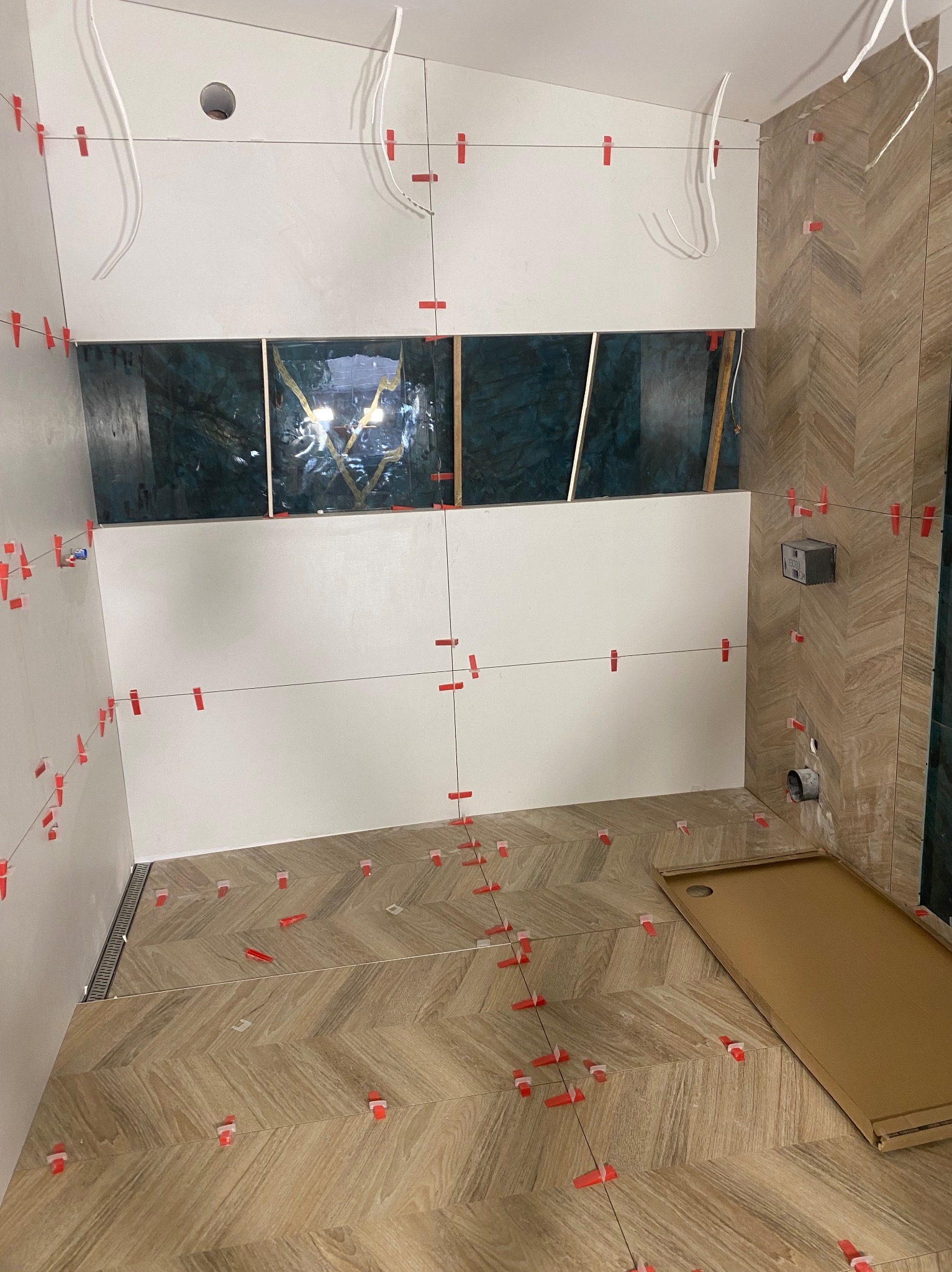 Łazienka w trakcie prac: ułożona podłoga w jodełkę, białe ściany z poziomymi liniami i czerwonymi klipsami, fragment zielonej ściany. Widoczne kable i otwory.