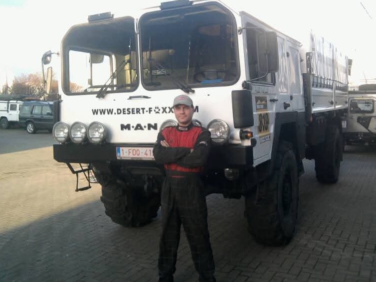 Kierowca w kombinezonie rajdowym stoi przed białym, terenowym MAN-em z napisem 'DESERT-F...XX' i numerem 810, gotowym do transportu w trudnych warunkach.