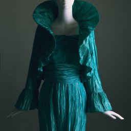 Adam Neubauer FOTOGRAFIA - Turkusowa, plisowana suknia z szerokim kołnierzem na manekinie, oświetlona studyjnie. Materiał ma wyraźną fakturę, a tło jest ciemnoszare.