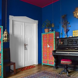Adam Neubauer FOTOGRAFIA - Kolorowe wnętrze pokoju z pianinem, regałem na książki i szafką. Intensywne barwy ścian i mebli w stylu boho. Dekoracje, lampy i dywan tworzą eklektyczną całość.