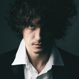 Adam Neubauer FOTOGRAFIA - Mężczyzna w półmroku, z rozwianymi włosami i spojrzeniem skierowanym wprost, ubrany w białą koszulę i czarną marynarkę, na ciemnym tle. Portret w stylu dramatycznym.