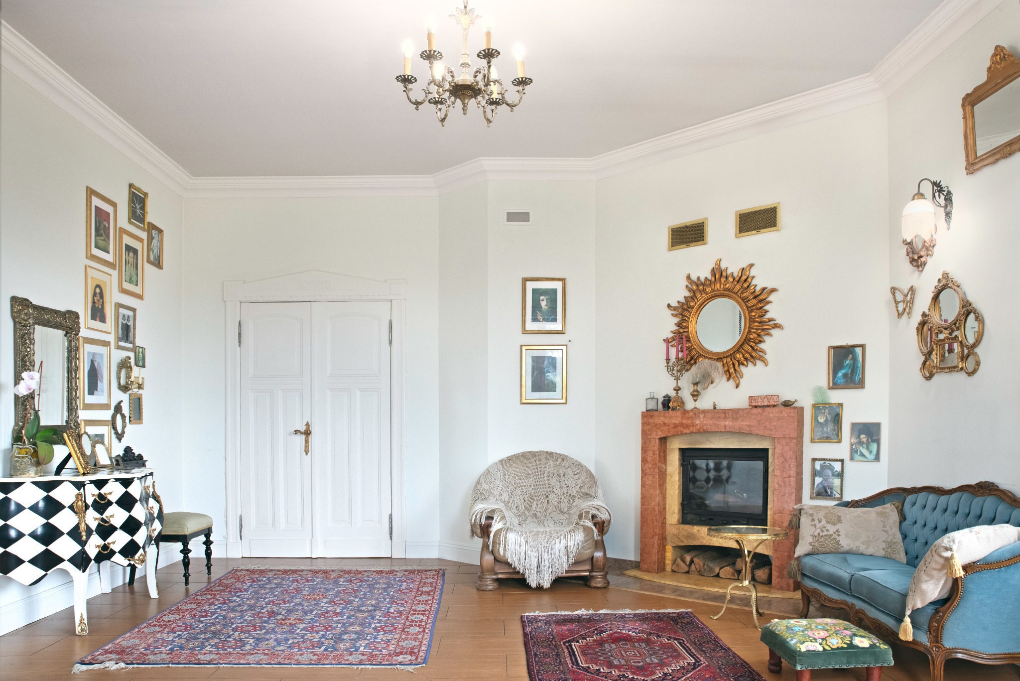 Salon w stylu retro z kominkiem, turkusową sofą i lustrami w złotych ramach. Na ścianach wiszą obrazy, a podłogę zdobią orientalne dywany. Centralnie umieszczony fotel z narzutą.