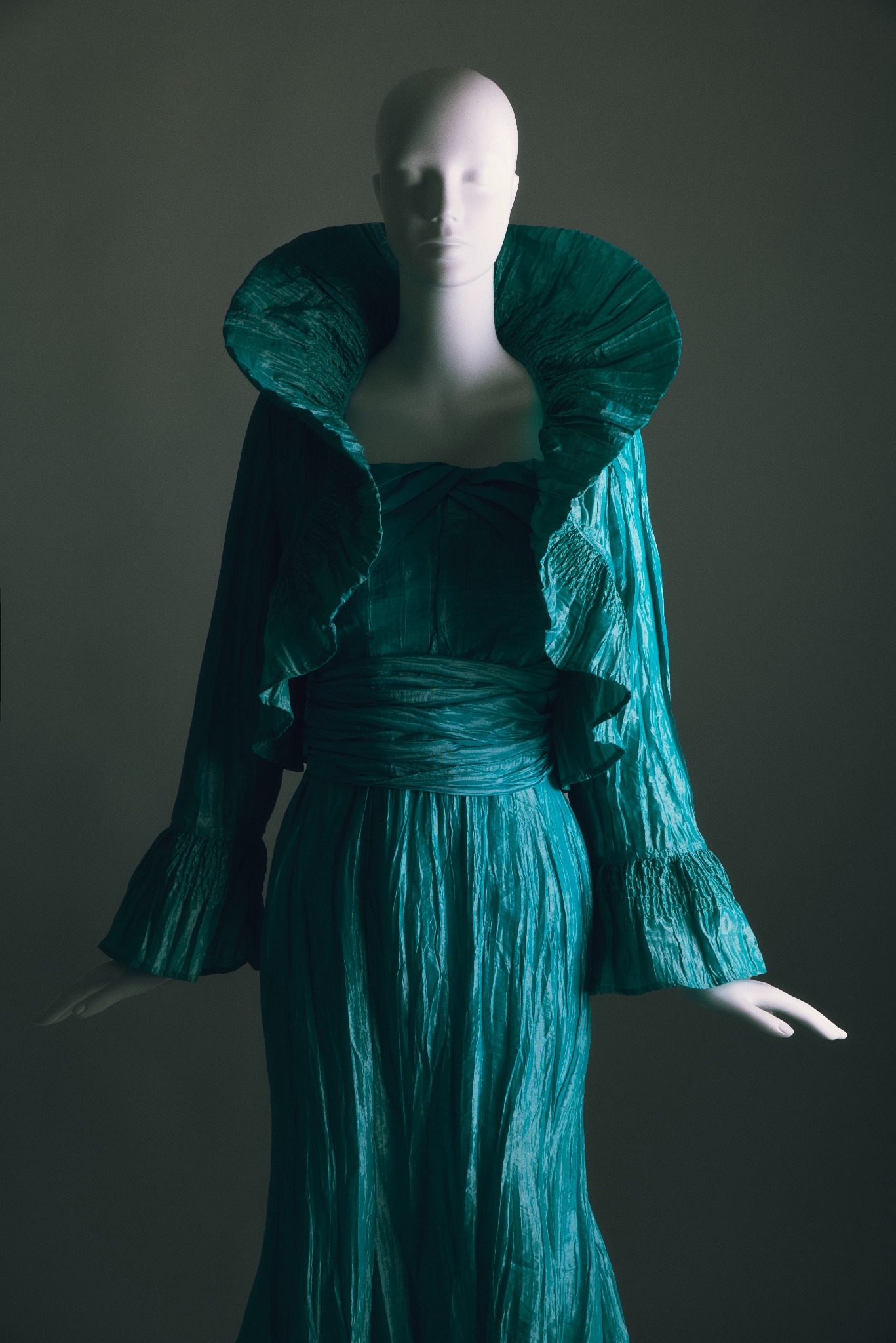 Turkusowa, plisowana suknia z szerokim kołnierzem na manekinie, oświetlona studyjnie. Materiał ma wyraźną fakturę, a tło jest ciemnoszare.