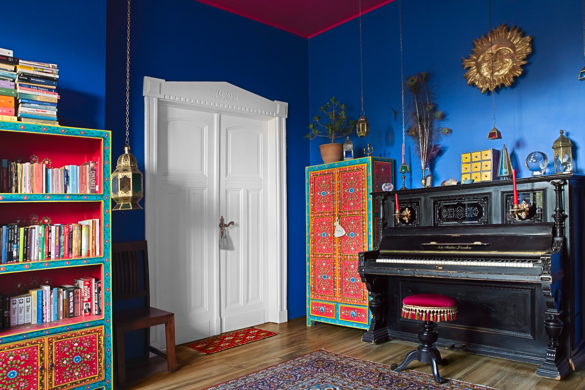 Kolorowe wnętrze pokoju z pianinem, regałem na książki i szafką. Intensywne barwy ścian i mebli w stylu boho. Dekoracje, lampy i dywan tworzą eklektyczną całość.
