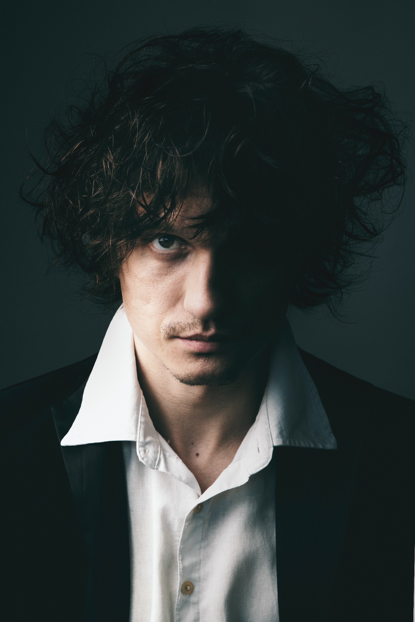 Mężczyzna w półmroku, z rozwianymi włosami i spojrzeniem skierowanym wprost, ubrany w białą koszulę i czarną marynarkę, na ciemnym tle. Portret w stylu dramatycznym.