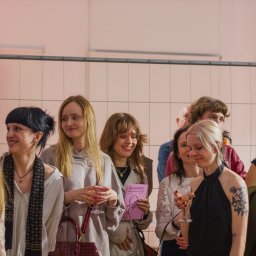 Danylo Vasiur - Grupa młodych osób, w większości kobiet, w pomieszczeniu z różowym oświetleniem. Jedna z kobiet trzyma w ręku różową książkę. Widoczna metalowa siatka w tle.