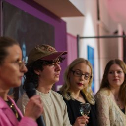 Danylo Vasiur - Grupa osób na evencie, kobieta w różowej marynarce z mikrofonem, w tle fioletowa ściana i obraz. Osoby w różnym wieku, z różnymi fryzurami i stylami ubioru.