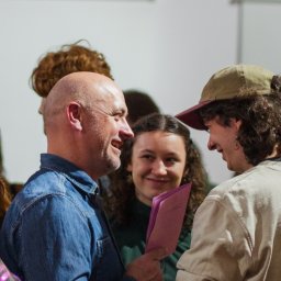 Danylo Vasiur - Uśmiechnięty mężczyzna w dżinsowej koszuli rozmawia z młodym mężczyzną w czapce, w tle grupa osób i biała ściana. W ręku trzyma różowe dokumenty.