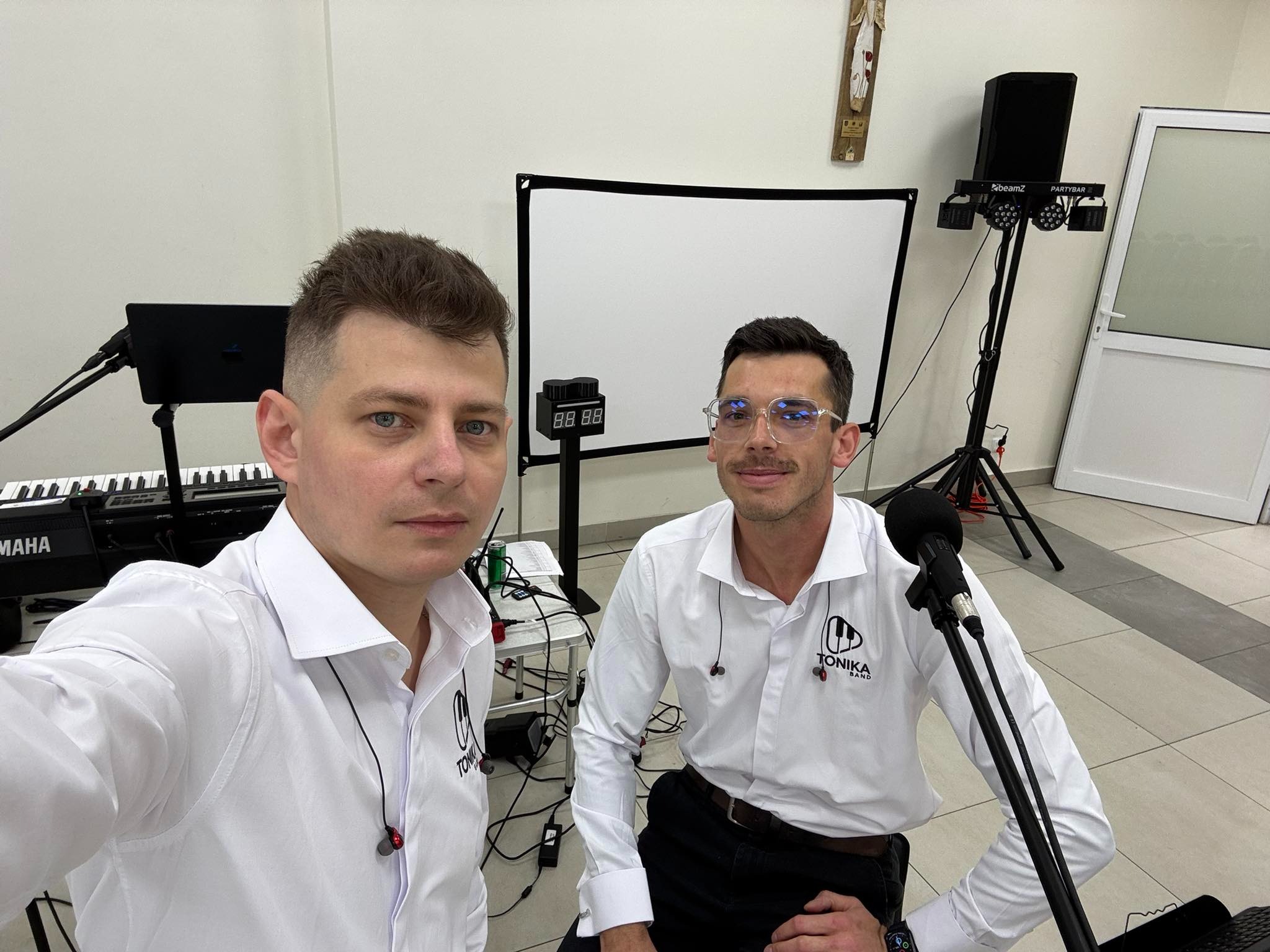 Dwóch muzyków zespołu Tonika Band w białych koszulach, z mikrofonami i sprzętem nagłośnieniowym w tle, gotowych do występu. Widoczny keyboard i oświetlenie.