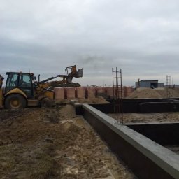 JG-BUD - Wylane fundamenty domu jednorodzinnego z wystającymi prętami zbrojeniowymi. W tle koparka transportuje materiały budowlane na placu budowy w pochmurny dzień.