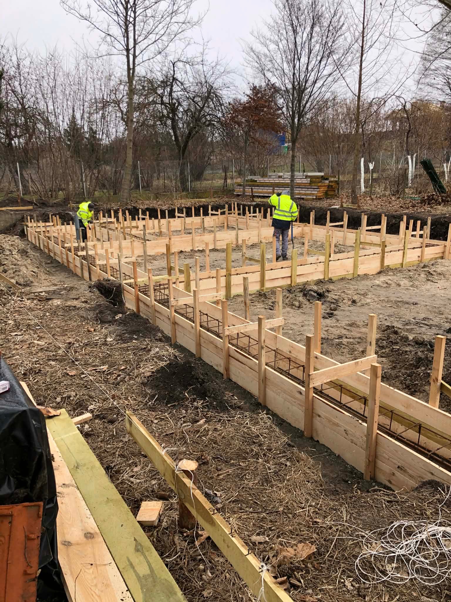 Zbrojenie i szalunek pod fundament. Dwóch pracowników w odblaskowych kamizelkach. Widoczne drewniane elementy konstrukcji i stalowe pręty. Tło: drzewa bez liści.