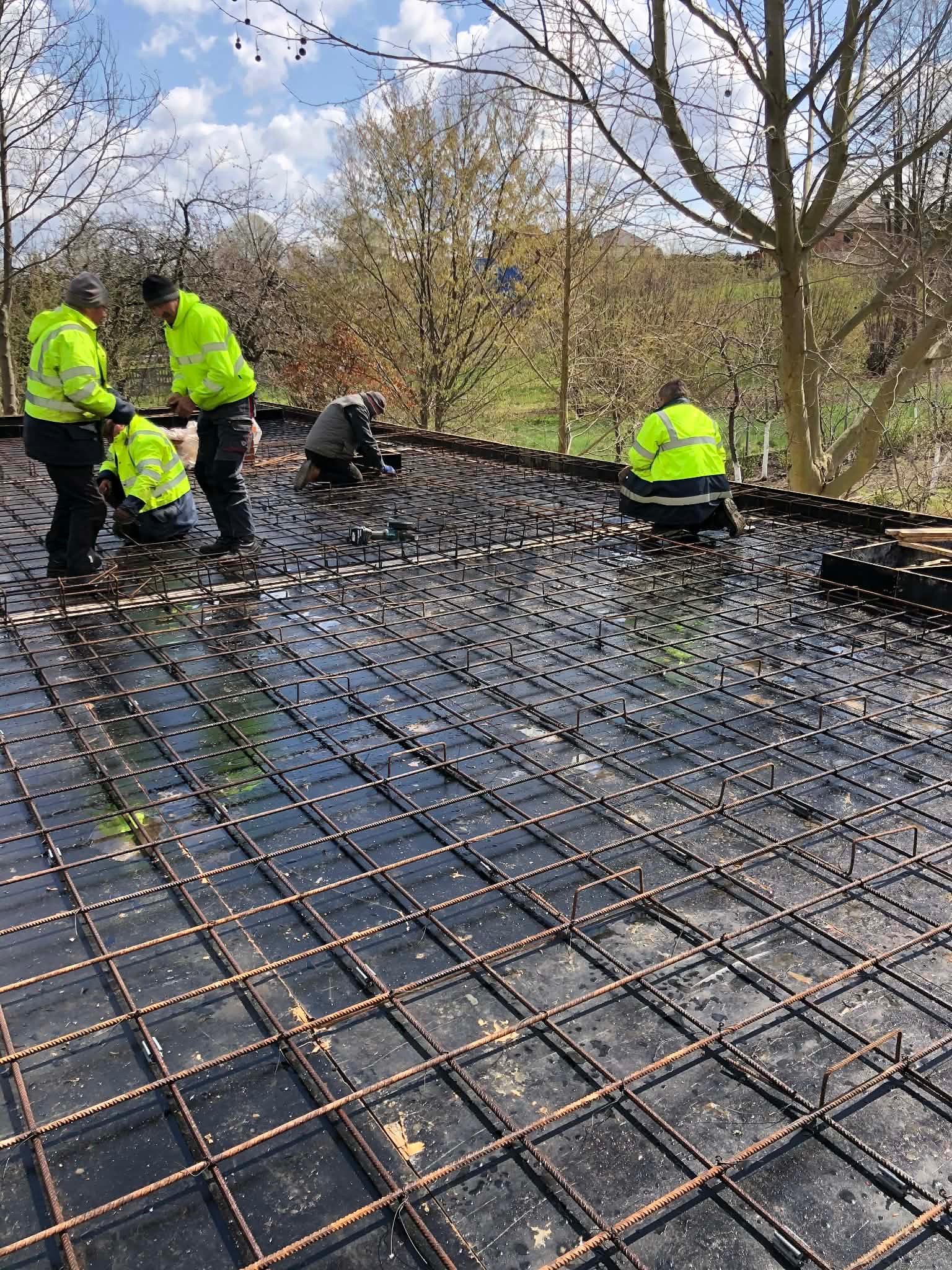 Prace zbrojarskie na budowie: robotnicy w odblaskowych kurtkach układają siatkę zbrojeniową na czarnej folii, przygotowując fundament pod budynek. Widok z góry.