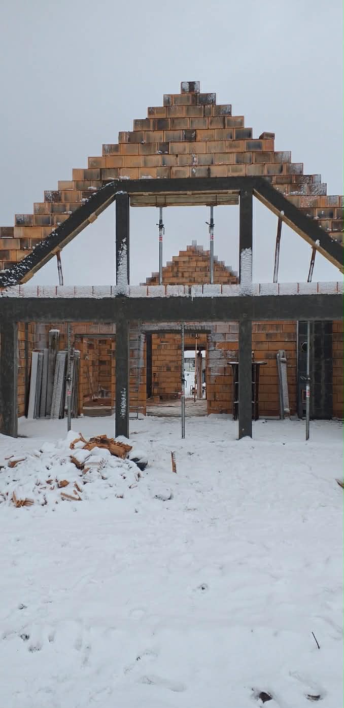 Surowy stan domu z cegły ceramicznej w zimowej scenerii. Widoczne metalowe podpory i śnieg na murach. Budowa w trakcie, zarys przyszłego budynku.