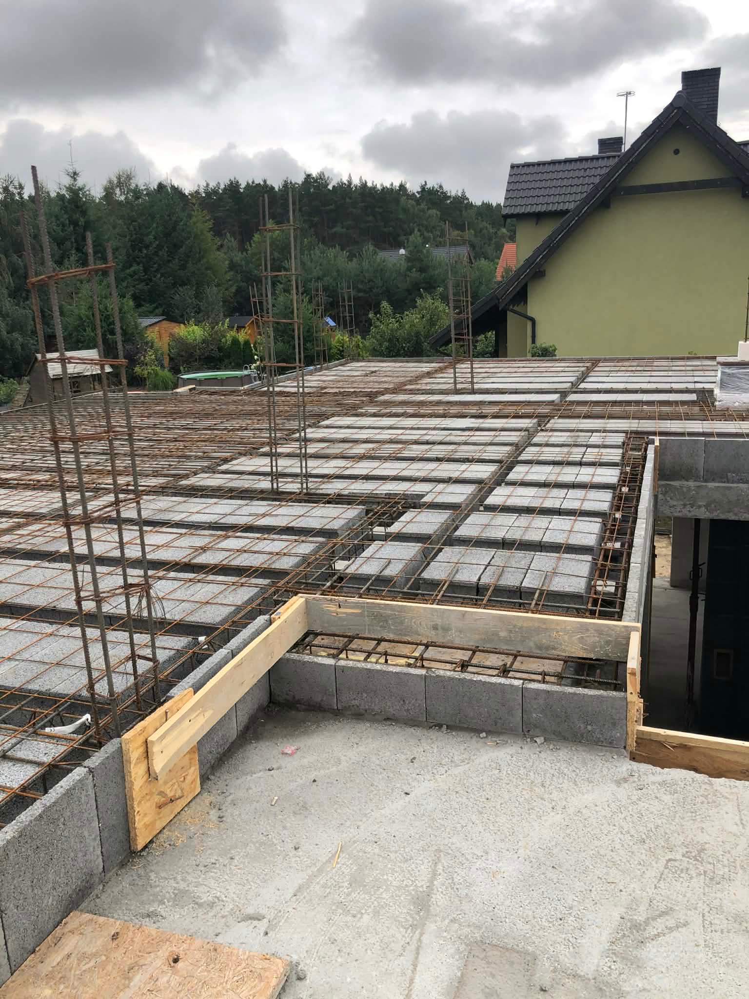 Zbrojenie stropu Teriva z betonowych pustaków szalunkowych, drewniane szalunki, widok na zalesione otoczenie i dom z ciemną dachówką w tle. Etap budowy domu.