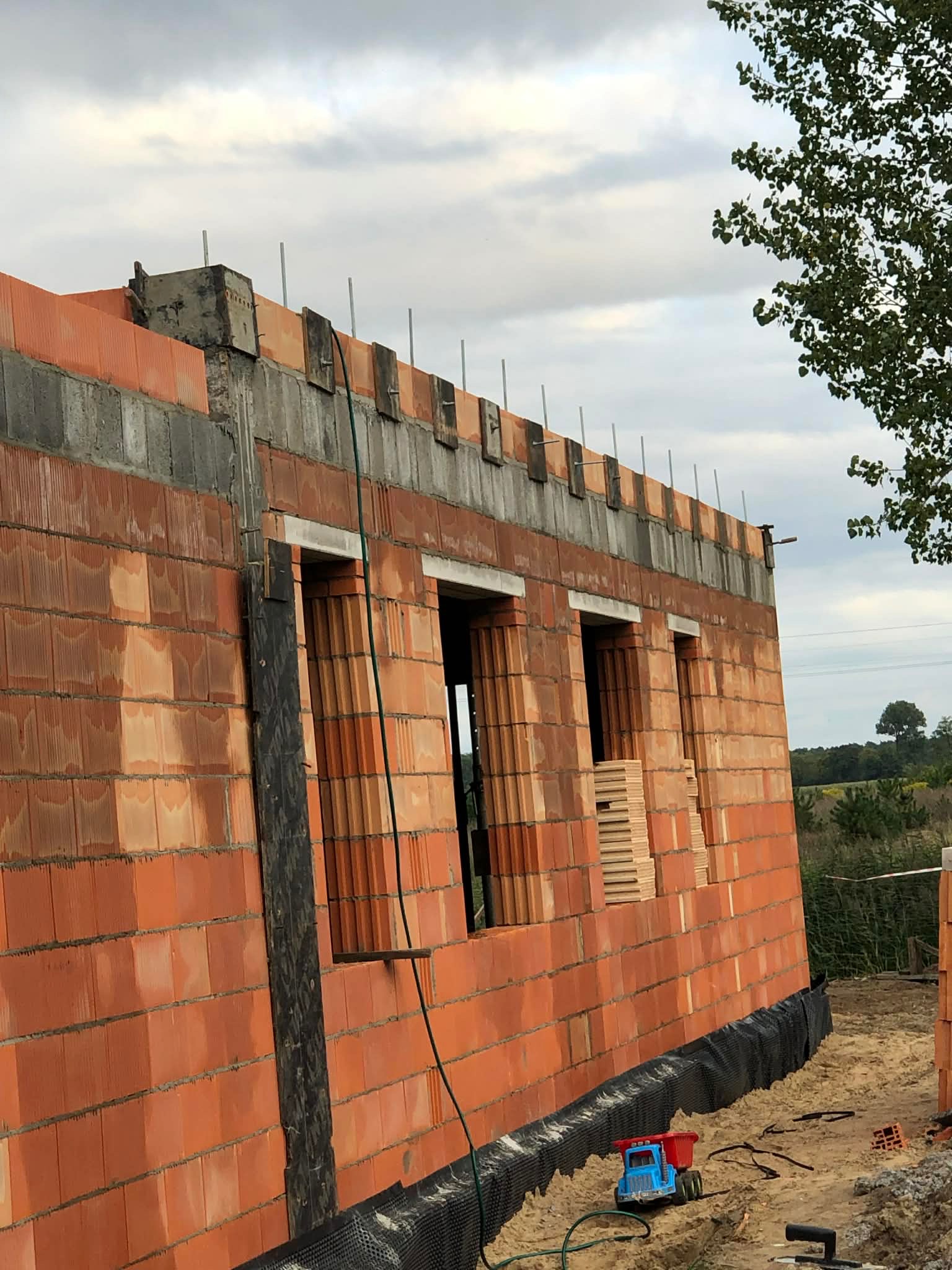 Wznoszenie murów z czerwonej cegły porotherm, widoczne otwory okienne i zbrojenie wieńca. Na dole zabawka - ciężarówka. Budowa w toku, tło: zielona łąka i pochmurne niebo.