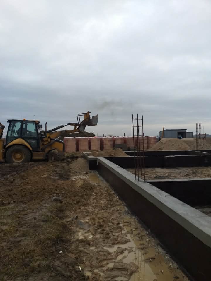 Wylane fundamenty domu jednorodzinnego z wystającymi prętami zbrojeniowymi. W tle koparka transportuje materiały budowlane na placu budowy w pochmurny dzień.