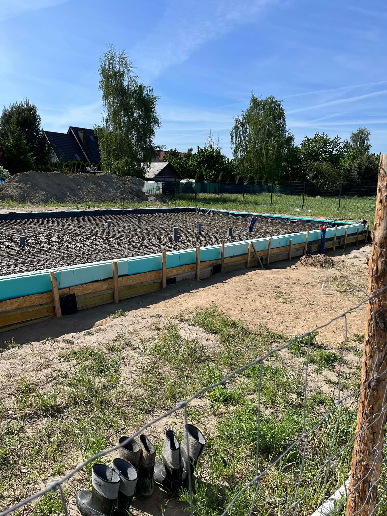 Zbrojenie fundamentu pod budowę domu jednorodzinnego, widoczne szalunki z izolacją termiczną, rury instalacyjne i siatka zbrojeniowa. W tle domy i drzewa.