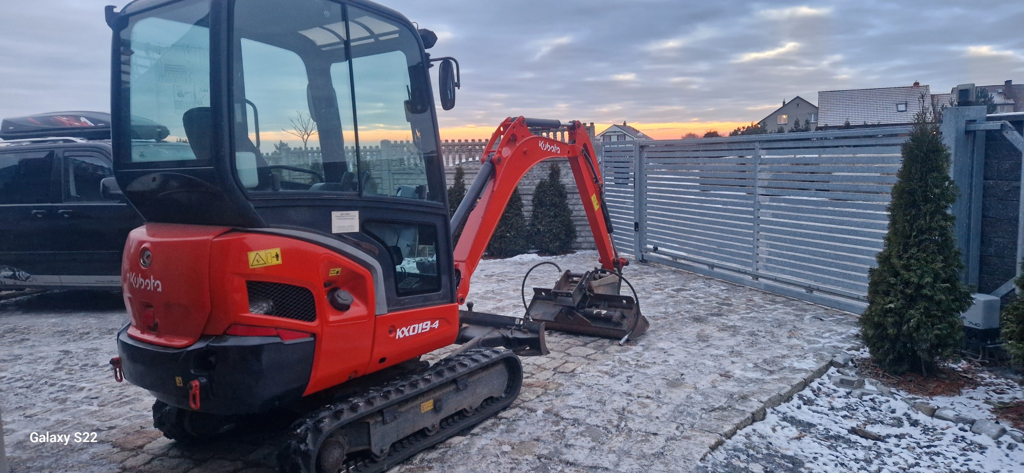 Pomarańczowa mini koparka Kubota KX019-4 na posesji z kostką brukową, w tle brama i zachodzące słońce. Widoczne ślady śniegu.