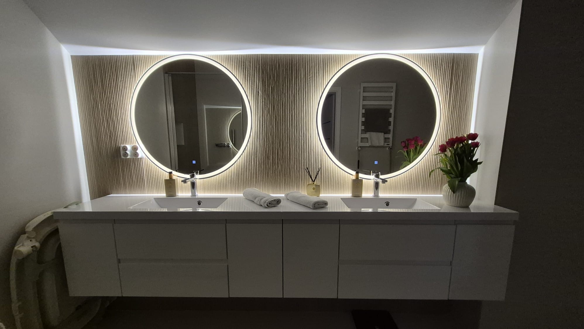 Nowoczesna łazienka z podwójną umywalką, okrągłymi lustrami z podświetleniem LED i dekoracyjną ścianą z pionowych paneli. Minimalistyczny design, kwiaty w wazonie.