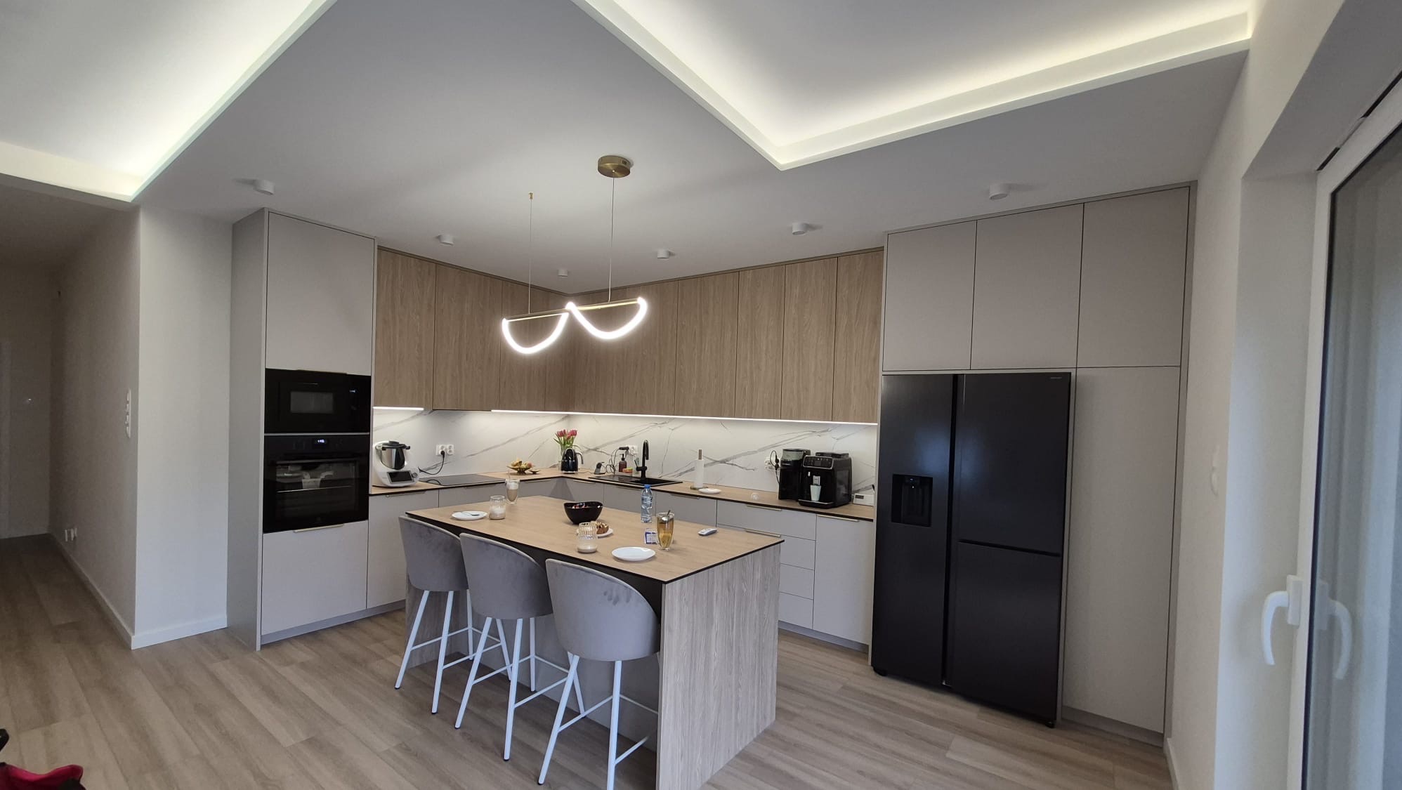 Nowoczesna kuchnia z wyspą i oświetleniem LED. Jasne drewno i szarość tworzą elegancką przestrzeń. Widoczny sprzęt AGD i detale aranżacji.