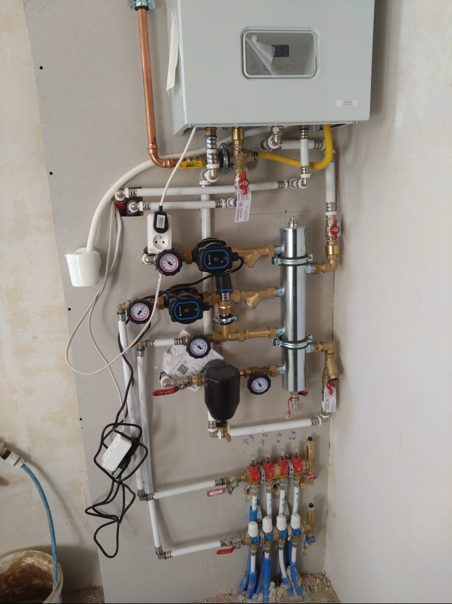 Nowoczesny piec gazowy z rozbudowaną instalacją hydrauliczną, widoczne liczne zawory, pompy obiegowe i manometry. Instalacja zamontowana na ścianie z płyt gipsowych.