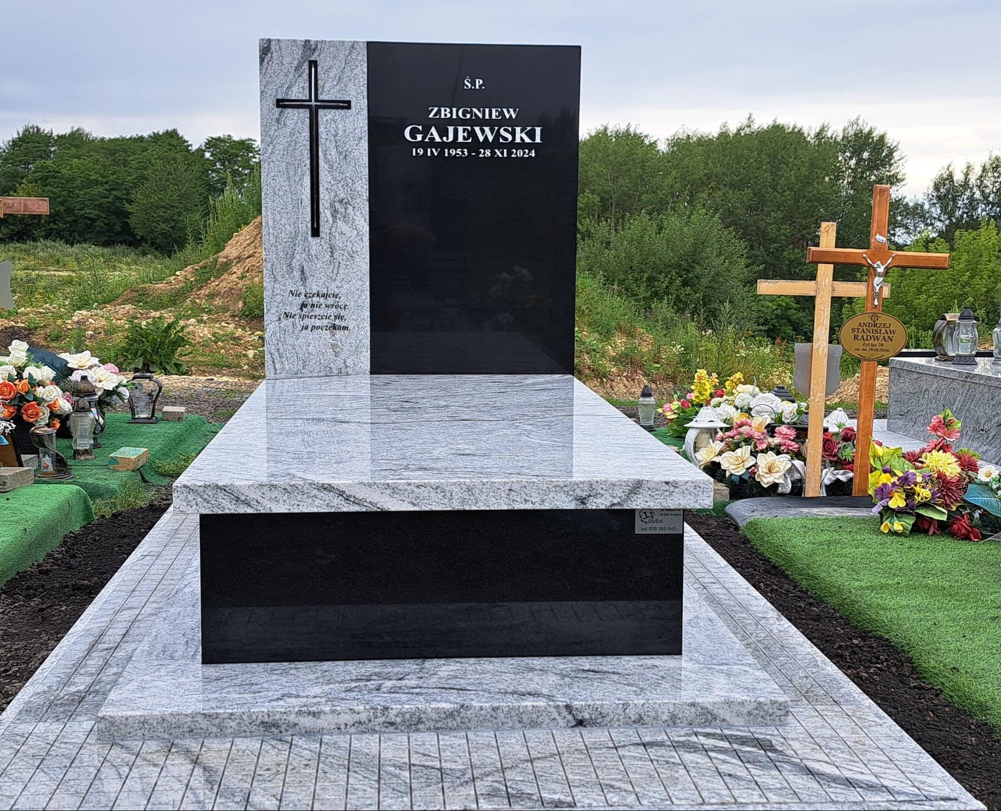 Granitowy nagrobek z czarną tablicą i wyrytym krzyżem, otoczony zielenią cmentarza. Jasnoszary kamień z delikatnym wzorem żyłkowania. Widoczne kwiaty i krzyże w tle.