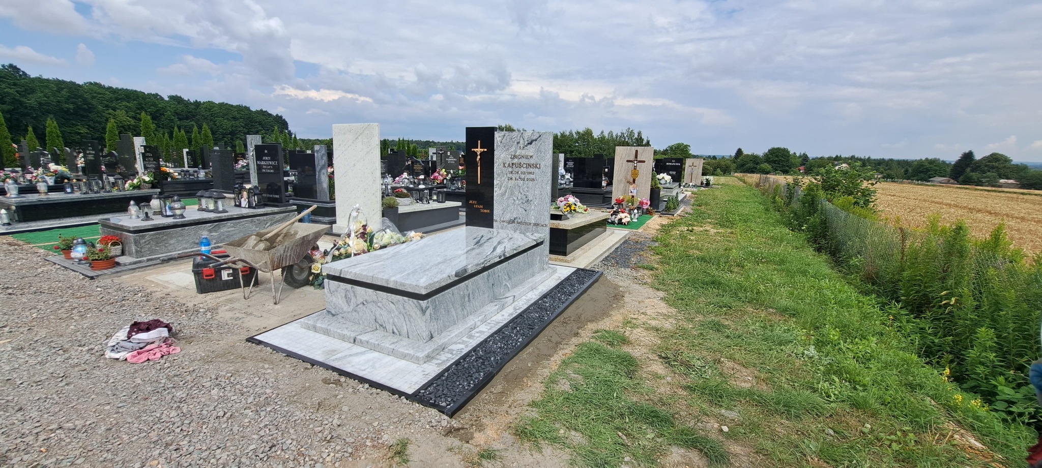 Jasny, marmurowy nagrobek z czarnymi akcentami na cmentarzu, otoczony zielenią i polami. Widok na nagrobki w perspektywie, pochmurne niebo.