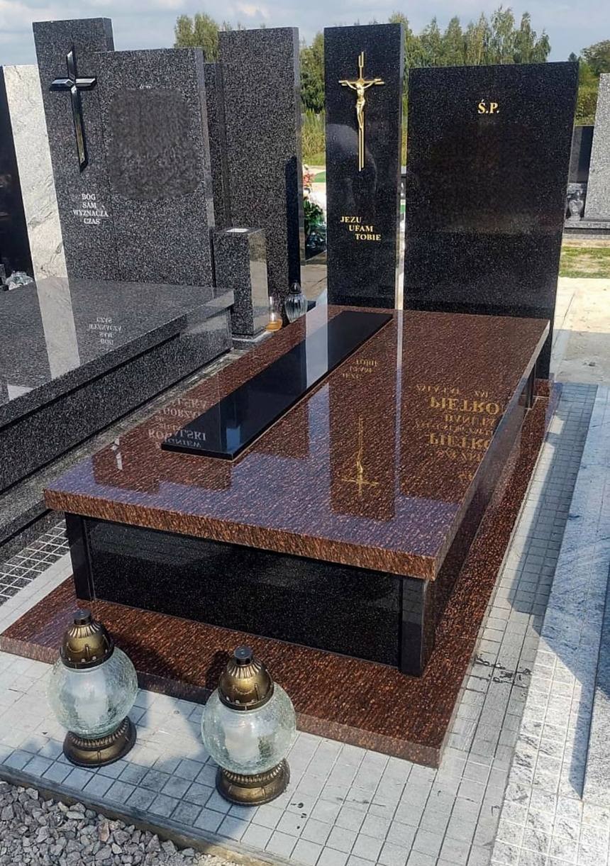 Brązowo-czarny, polerowany nagrobek z granitu z inskrypcjami i krzyżem, oświetlony naturalnym światłem, z lampionami na płycie chodnikowej.