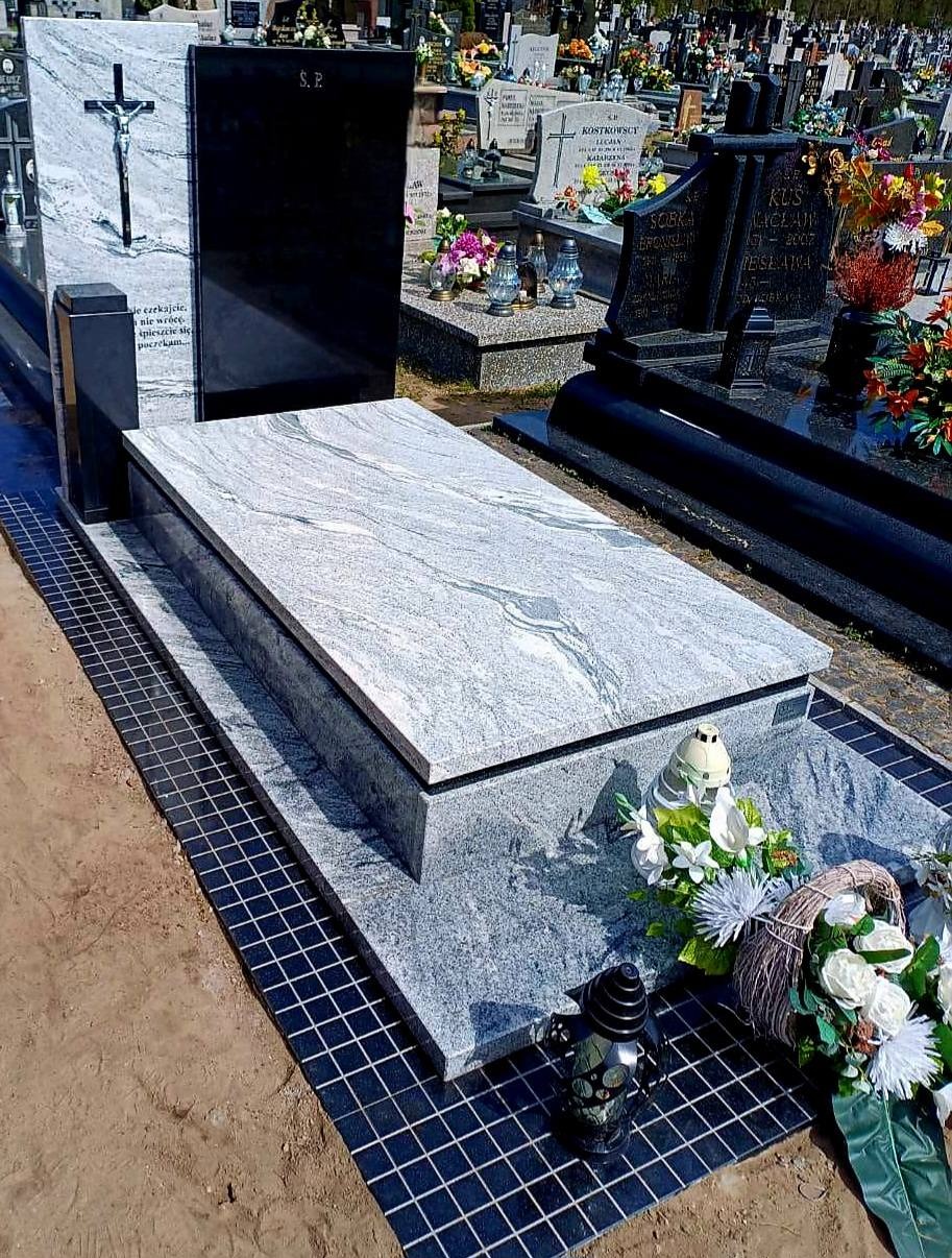 Granitowy nagrobek z jasnoszarym blatem o falistej fakturze, czarną tablicą i krzyżem, otoczony kwiatami i lampionami na cmentarzu. Widok z góry.