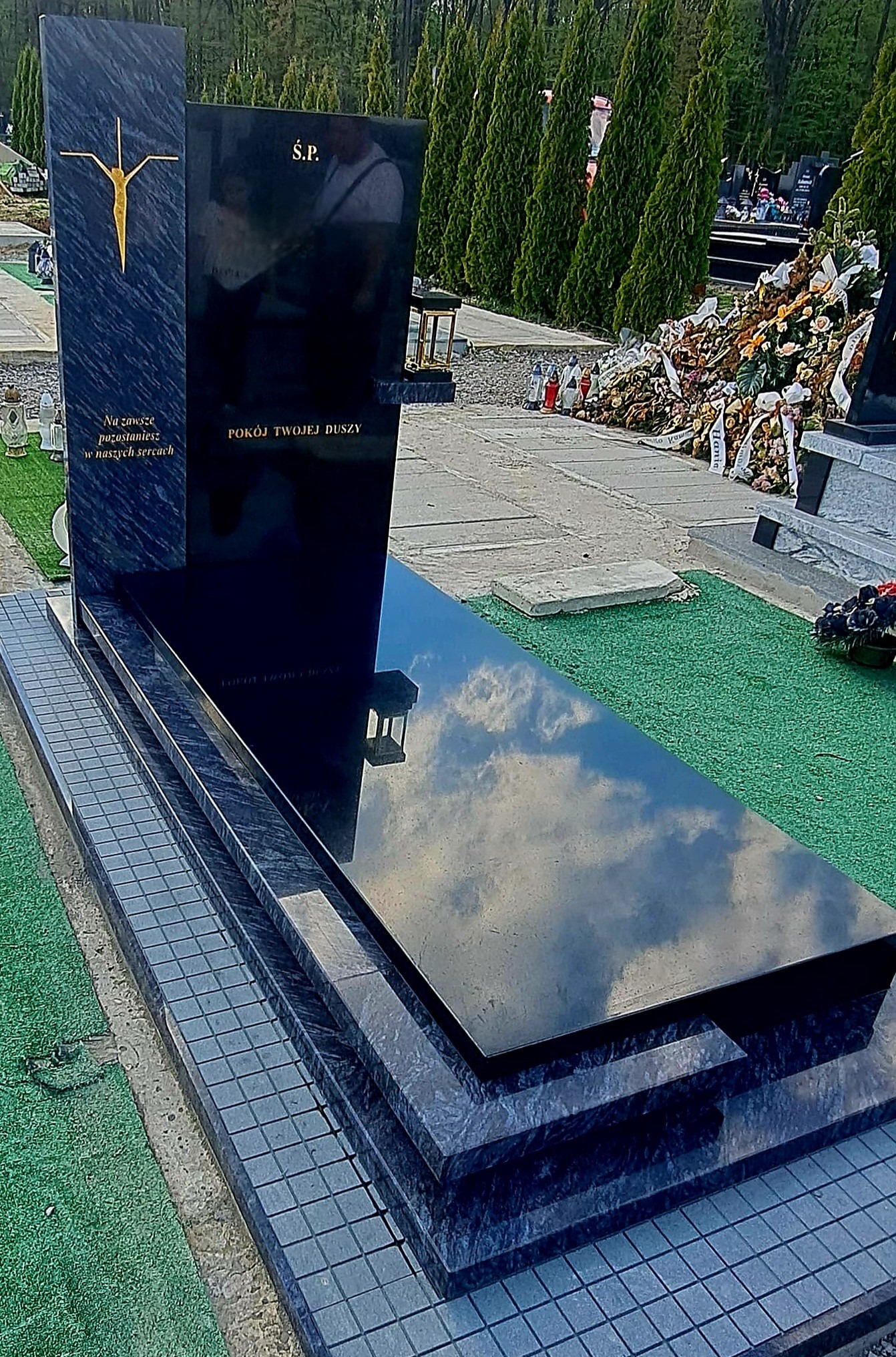 Czarny, polerowany nagrobek z krzyżem i napisem 'Ś.P. Pokój Twojej Duszy' na cmentarzu, z odbiciem nieba w płycie. Widoczne kwiaty i drzewa w tle.