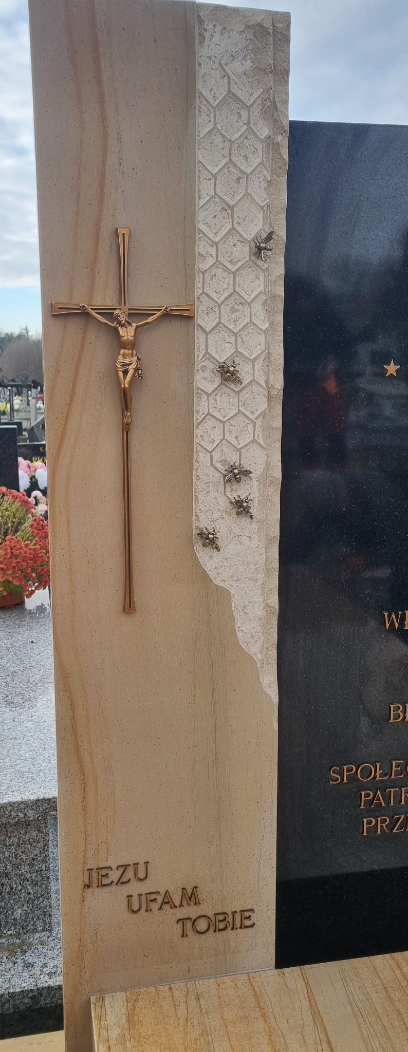 Beżowo-czarny nagrobek z motywem plastra miodu i figurkami pszczół. Na jasnej płycie napis 'Jezu ufam Tobie' i krucyfiks. Widok na cmentarz w tle.