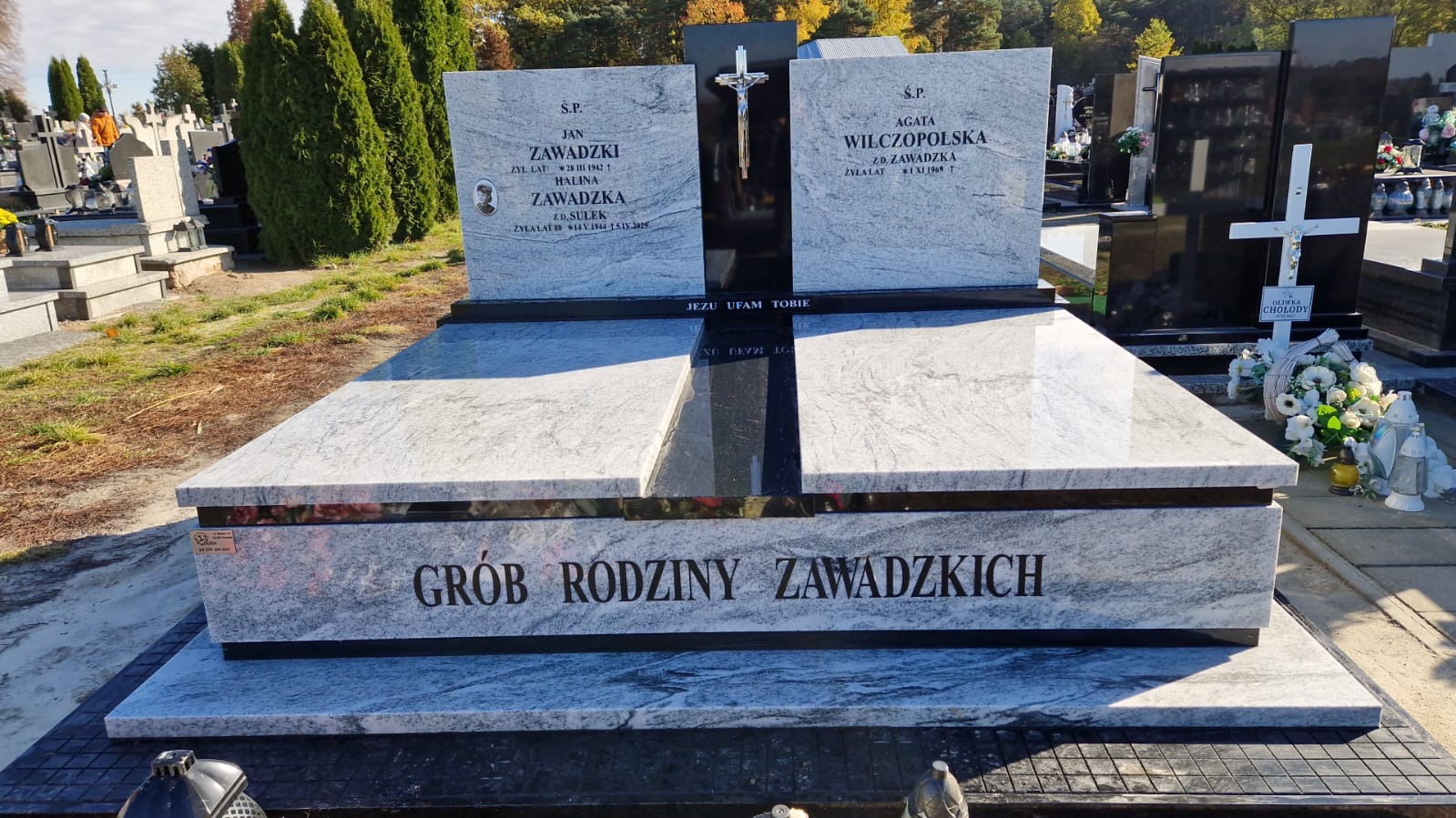 Granitowy nagrobek rodzinny z inskrypcjami i zdjęciami, otoczony zielenią cmentarza. Jasny kamień z ciemnymi akcentami, krzyż na górze. Napis: Grób Rodziny Zawadzkich.