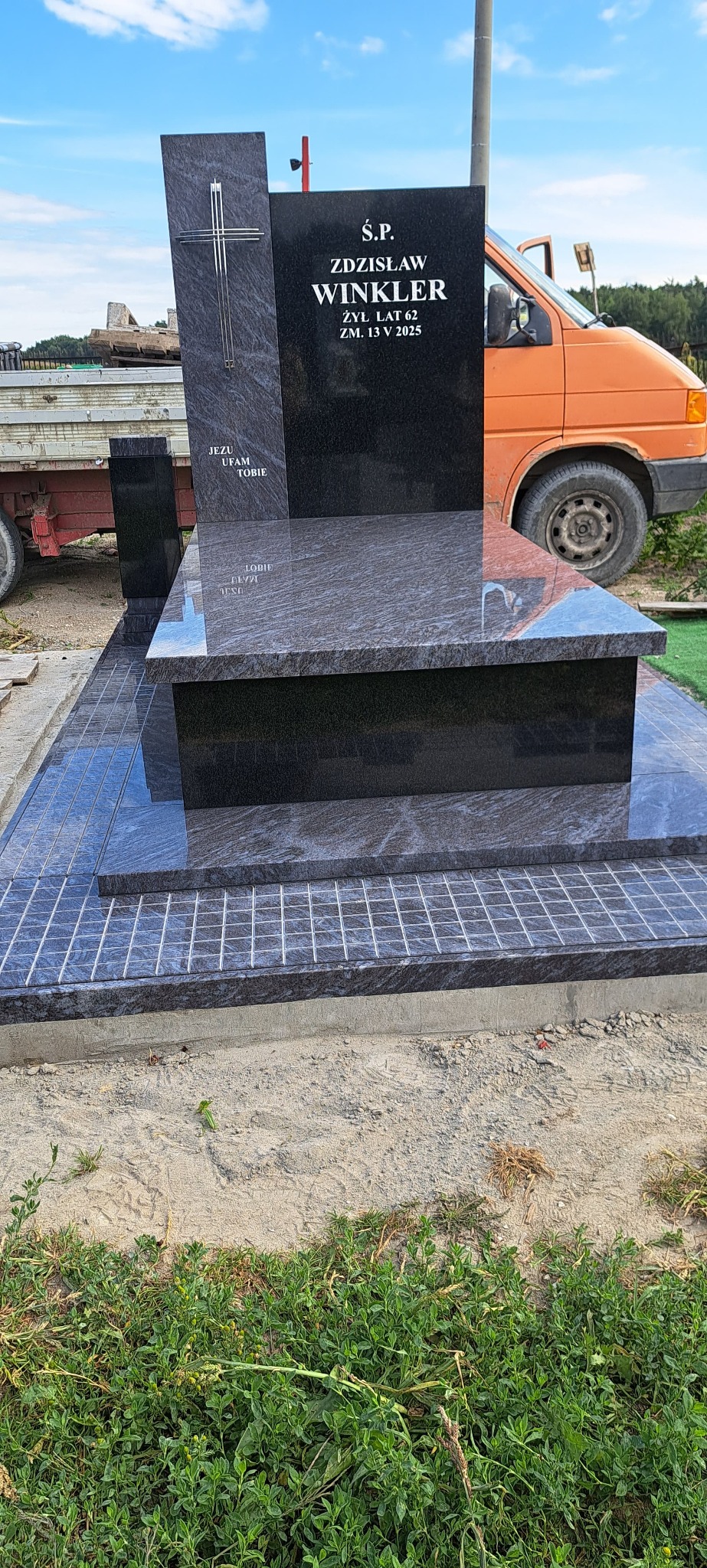 Granitowy nagrobek z inskrypcją 'Ś.P. Zdzisław Winkler', krzyżem i datą śmierci 13 V 2025, na tle pomarańczowego samochodu dostawczego i zieleni.