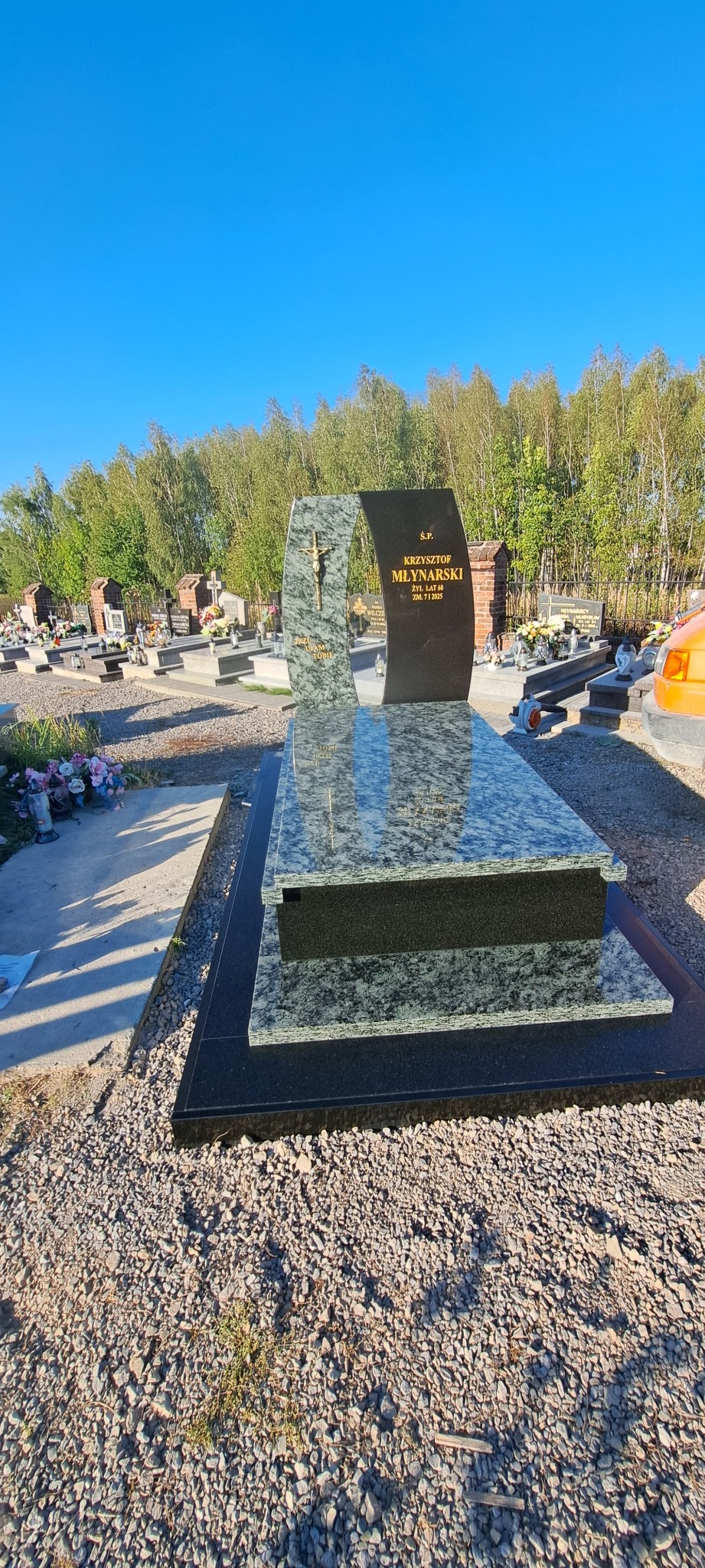 Nowoczesny nagrobek z jasnego granitu z krzyżem i ciemną tablicą inskrypcyjną na cmentarzu, w tle drzewa i inne groby, słoneczny dzień.